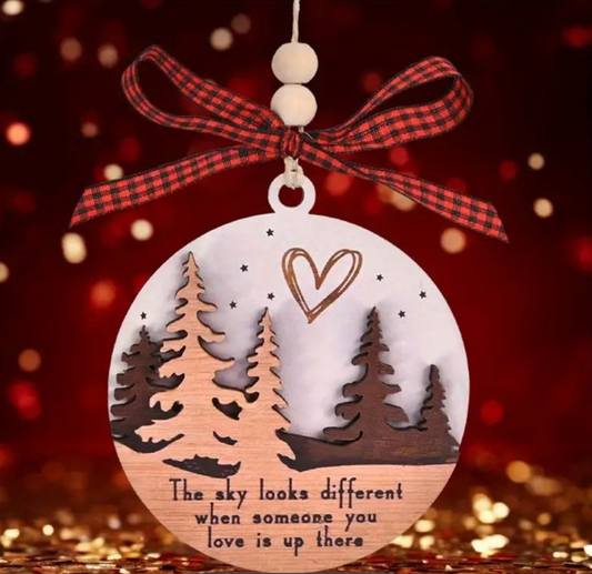 a round wooden ornament with engraved trees, a heart, and the text 'The sky looks different when someone you love is up there.' The ornament is decorated with a red plaid ribbon and a wooden bead, against a blurred festive background