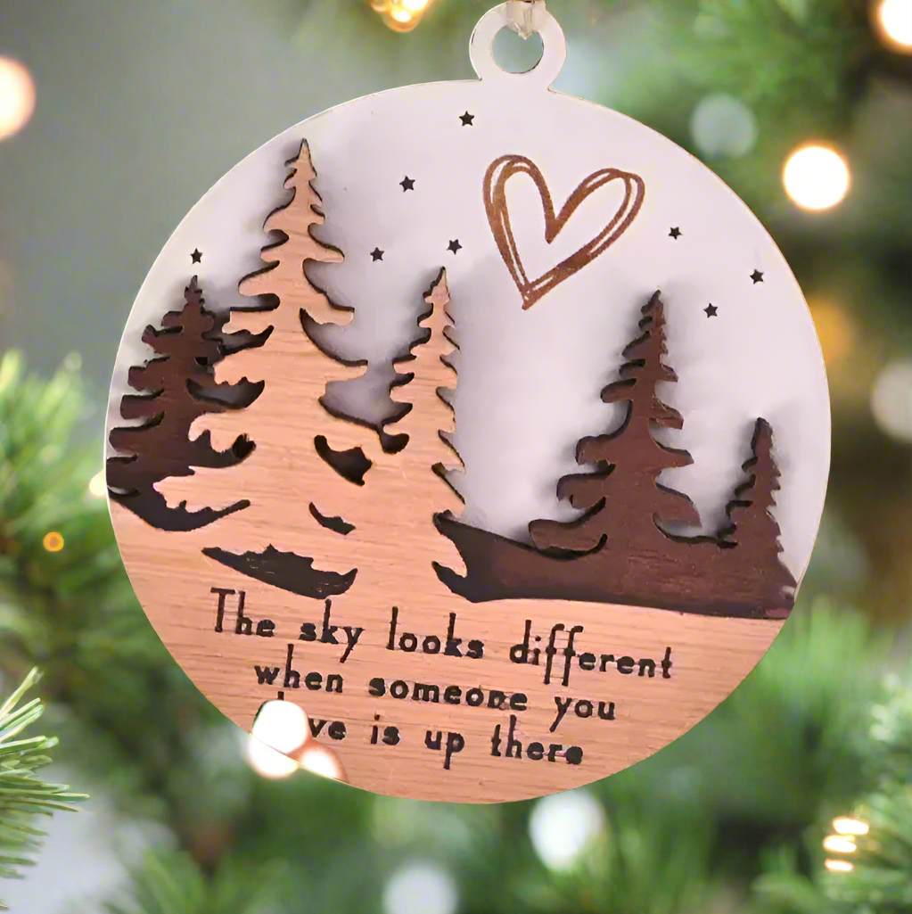 Handsholding a round wooden ornament with engraved trees, a heart, and the text 'The sky looks different when someone you love is up there.' The ornament is decorated with a red plaid ribbon and a wooden bead, against a blurred festive background